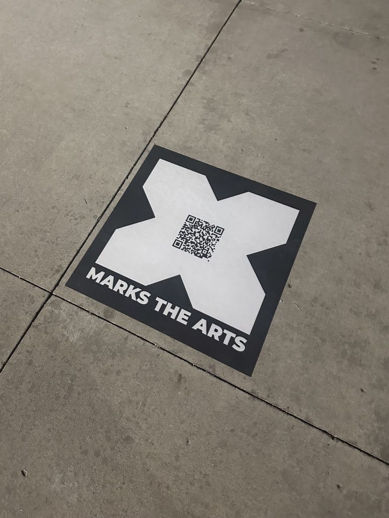 A “X Marks the Arts sidewalk sign directs summer vacationers and locals to the arts, culture, and entertainment venues throughout downtown Iowa City.